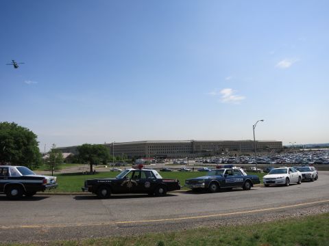 1991 Ford LTD Crown Victoria - line of vintage police cruisers is parked along a road in front of the Pentagon as a helicopter flies in the blue sky above.