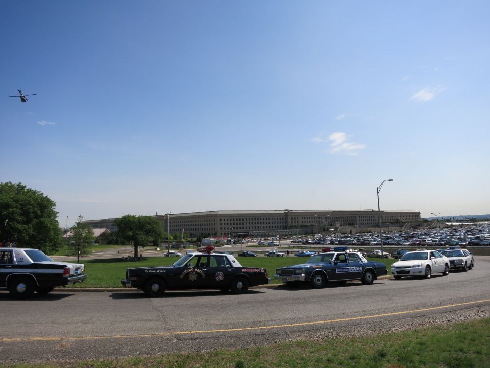 Row of vintage police cars parked by the Pentagon with a helicopter flying overhead