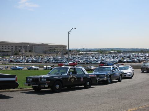 1991 Ford LTD Crown Victoria - black Highway Patrol sedan leads a line of vintage police cruisers parked beside a vast lot in front of a large government building.