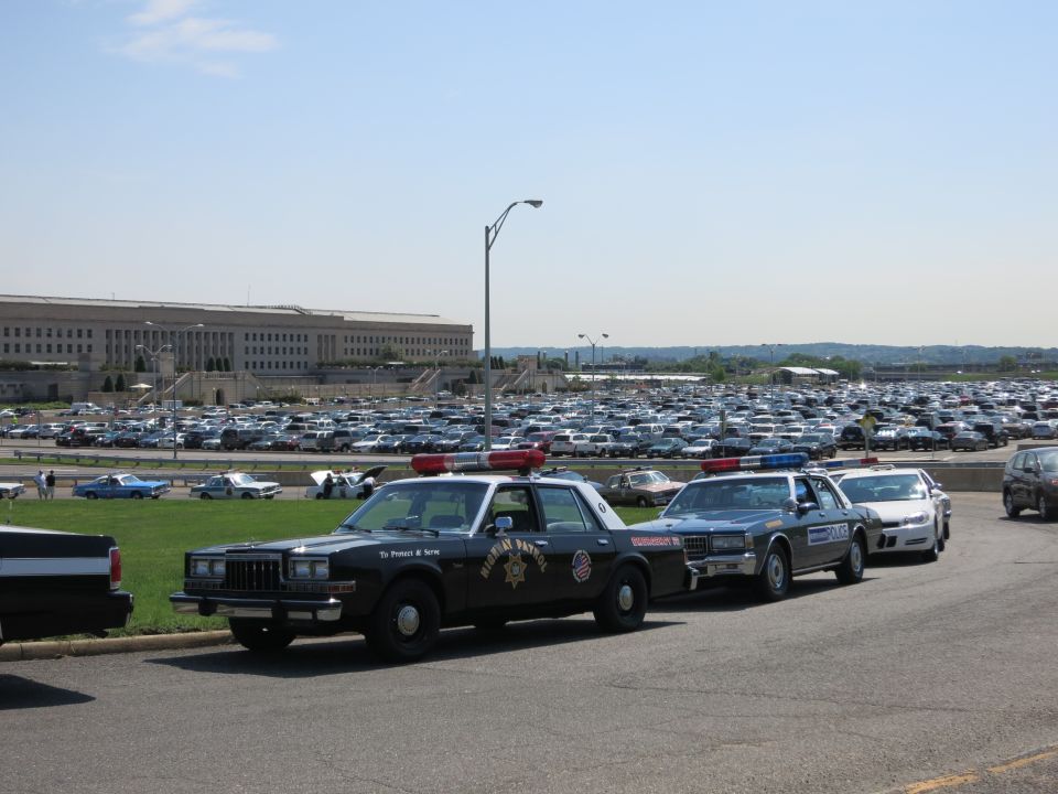 Black Highway Patrol sedan and other police cars lined up near a large government building and packed parking lot