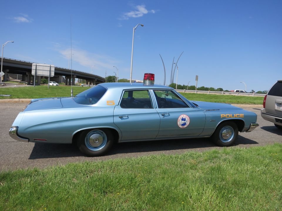 Light blue four-door Police sedan parked on grass beside a highway, side profile