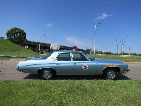 1991 Ford LTD Crown Victoria - Side profile of a light blue four-door police sedan marked POLICE parked on pavement beside a grassy embankment and overpass.