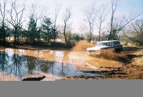 1978 Ford Bronco - A 1978 Ford Bronco navigates a muddy obstacle, kicking up a significant spray.