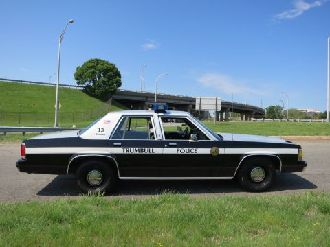 1991 Ford LTD Crown Victoria - black-and-white sedan lettered TRUMBULL POLICE sits in profile on pavement near a highway overpass.
