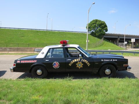 1991 Ford LTD Crown Victoria - black-and-white sedan marked HIGHWAY PATROL with a star shield decal sits in profile beside a guardrail and overpass.