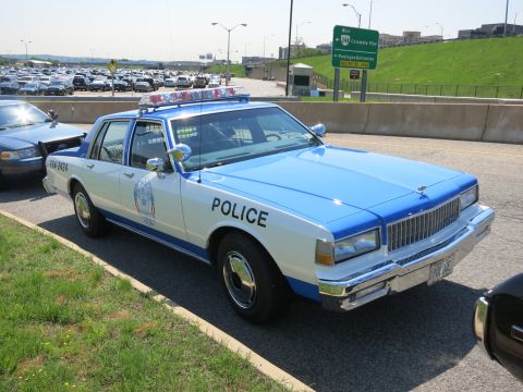1991 Ford LTD Crown Victoria - white-and-blue police sedan marked POLICE with a roof light bar is parked near a green highway sign and a full parking lot.