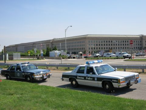 1991 Ford LTD Crown Victoria - dark State Police cruiser and a white Boston Police cruiser are parked on a road before a long stone government building.