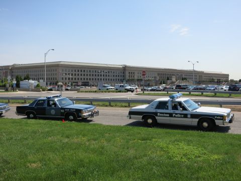1991 Ford LTD Crown Victoria - dark State Police cruiser and a white Boston Police cruiser are parked on a grassy strip before a long government building.
