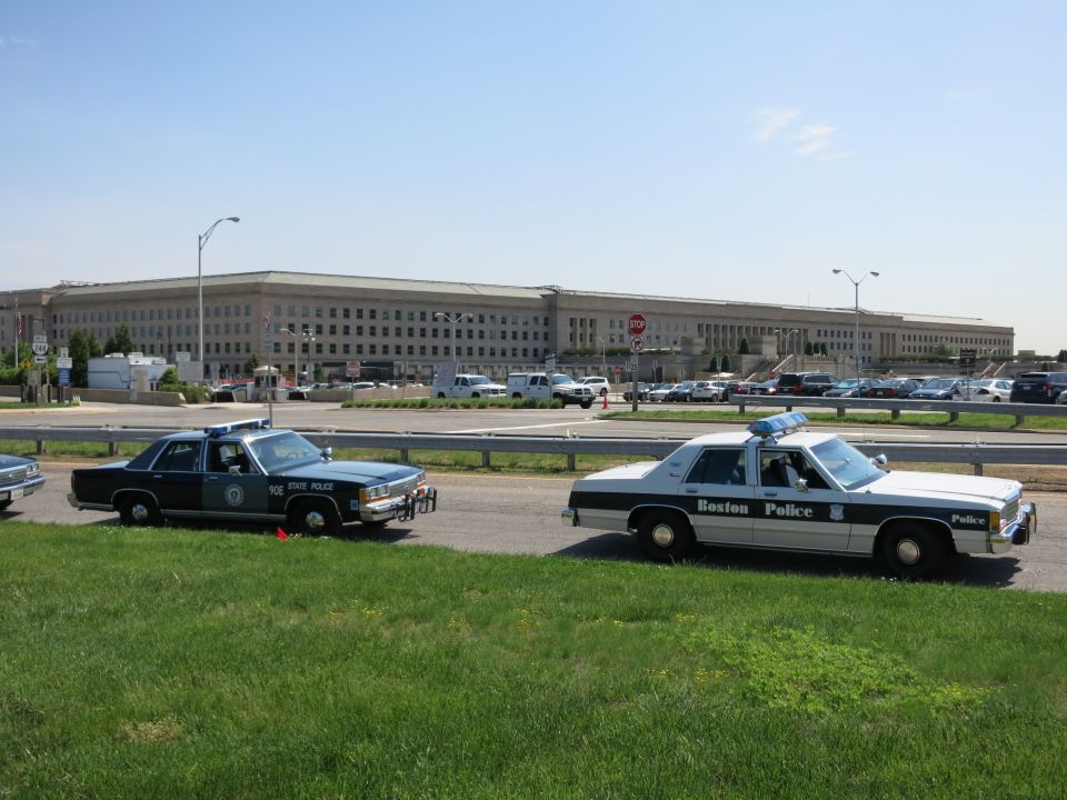 Dark State Police cruiser and white Boston Police cruiser parked on grass by a government building