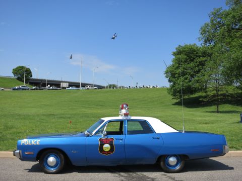 1991 Ford LTD Crown Victoria - blue-and-white police sedan marked POLICE with roof light and tall whip antenna sits on grass as helicopters fly overhead.