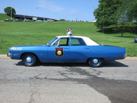 1991 Ford LTD Crown Victoria - Driver-side profile of a blue police sedan with white roof, gold shield decal, roof light and whip antenna, parked on pavement.
