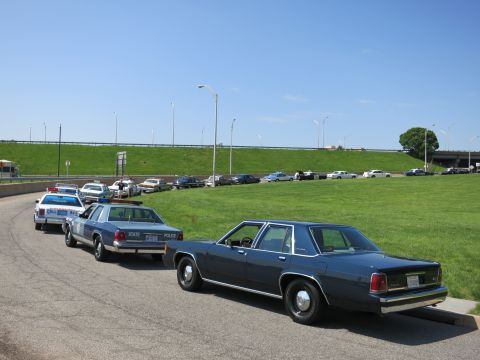 1991 Ford LTD Crown Victoria - row of vintage sedans, including State Police cruisers, parked along a curving road beside a grassy embankment.