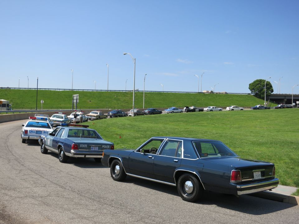 Several vintage sedans including State Police cruisers parked along a curving road by a grassy slope