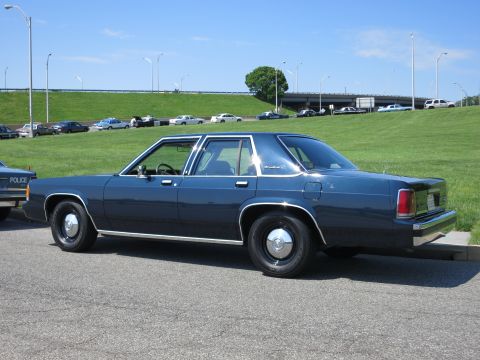1991 Ford LTD Crown Victoria - Rear three-quarter view of a dark blue four-door sedan parked on pavement near a marked police car and a highway overpass.