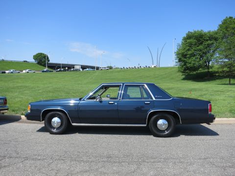 1991 Ford LTD Crown Victoria - Side profile of a dark blue four-door sedan with black steel wheels, parked on pavement before a grassy slope and overpass.