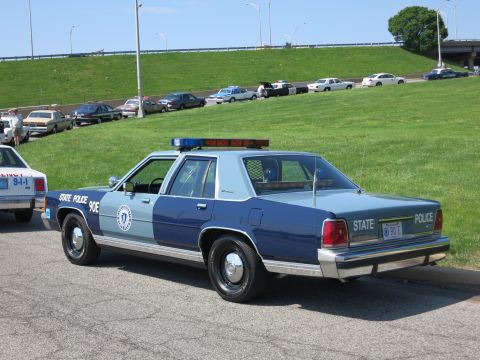 1991 Ford LTD Crown Victoria - blue-and-white STATE POLICE cruiser with roof light bar sits in rear three-quarter view near other parked cruisers and an overpass.