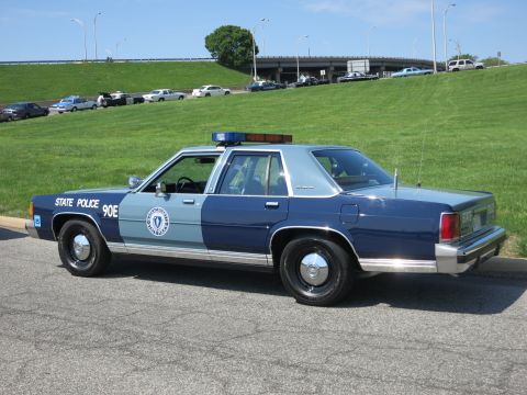 1991 Ford LTD Crown Victoria - blue-and-silver Massachusetts State Police sedan marked 90E sits parked on a road below a grassy embankment and highway overpass.