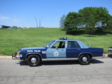 1991 Ford LTD Crown Victoria - blue-and-silver State Police sedan marked 90E is shown in profile, parked along a road with a grassy hillside behind it.