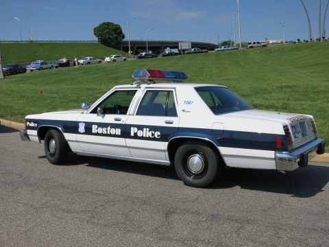 1991 Ford LTD Crown Victoria - white-and-blue Boston Police sedan marked 7087 sits parked on a road with a highway overpass and green hill in the background.