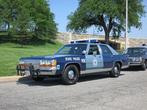 1991 Ford LTD Crown Victoria - blue-and-silver State Police sedan marked 90E, fitted with a front push bumper, parked in front of a stone overpass and trees.