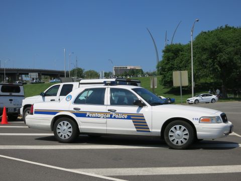 1991 Ford LTD Crown Victoria - white Pentagon Police sedan numbered 110 with blue and yellow striping and a roof lightbar, parked at a crosswalk.