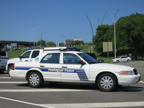 1991 Ford LTD Crown Victoria - white Pentagon Police sedan marked 110 with blue and yellow striping, parked near a road with a white SUV and highway behind.