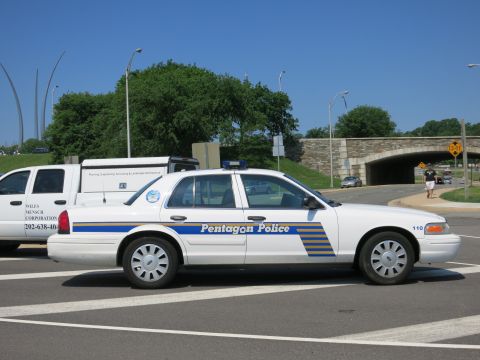 1991 Ford LTD Crown Victoria - white Pentagon Police sedan numbered 110 parked in profile next to a lettered work van, with a stone overpass behind.