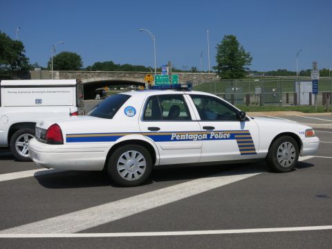 1991 Ford LTD Crown Victoria - white Pentagon Police sedan marked 110 with blue and yellow stripes, parked in profile near an overpass and a work van.