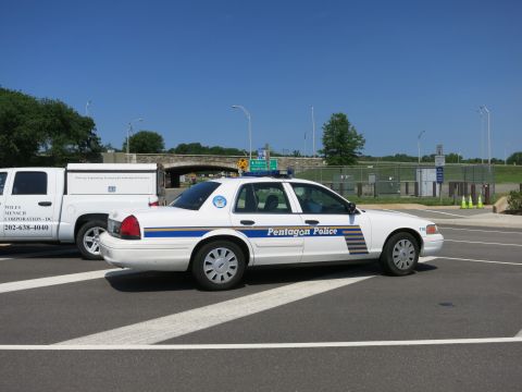 1991 Ford LTD Crown Victoria - white Pentagon Police sedan parked at an angle beside a lettered work van, with a stone overpass and fenced lot behind.