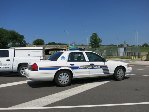 1991 Ford LTD Crown Victoria - white Pentagon Police sedan with blue and yellow stripes parked beside a work van, viewed from the rear near an overpass.