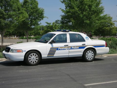 1991 Ford LTD Crown Victoria - white Pentagon Police sedan numbered 106 with blue and yellow striping and a lightbar, parked in a tree-lined lot.