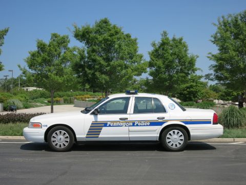 1991 Ford LTD Crown Victoria - white Pentagon Police sedan marked 106 shown in full profile, parked along a curb backed by green trees.