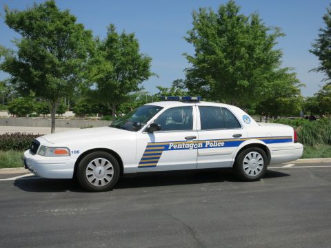 1991 Ford LTD Crown Victoria - white Pentagon Police sedan numbered 106 with blue and yellow stripes, parked in profile in front of leafy trees.