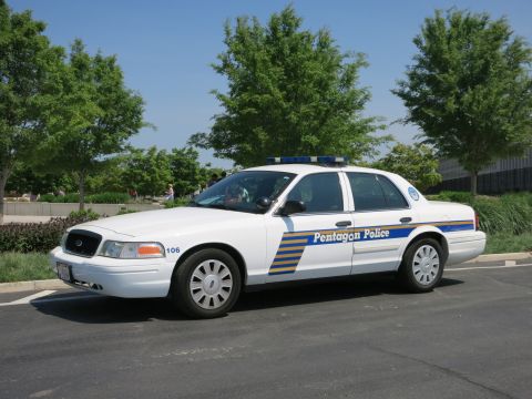 1991 Ford LTD Crown Victoria - white Pentagon Police sedan marked 106 with a roof lightbar, viewed from the front quarter in a tree-lined parking area.