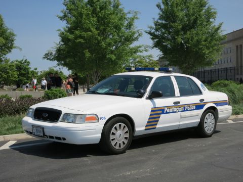 1991 Ford LTD Crown Victoria - white Ford Crown Victoria sedan lettered Pentagon Police, unit 106, parked on pavement with trees and bystanders behind.