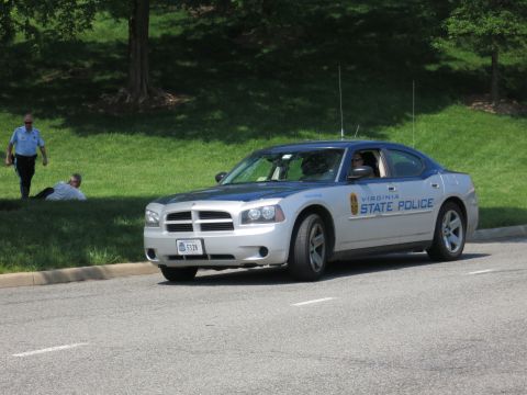1991 Ford LTD Crown Victoria - silver Dodge Charger sedan marked Virginia State Police moves along a curving road with grass and an officer in the background.