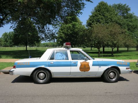 1991 Ford LTD Crown Victoria - white and blue police sedan, unit 401, with a gold door shield and roof beacon parked on a park road profile view.