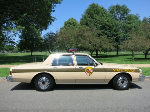 1991 Ford LTD Crown Victoria - Profile view of a tan State Police sedan with a Maryland-style gold shield and chrome wheels parked on a sunny park road.