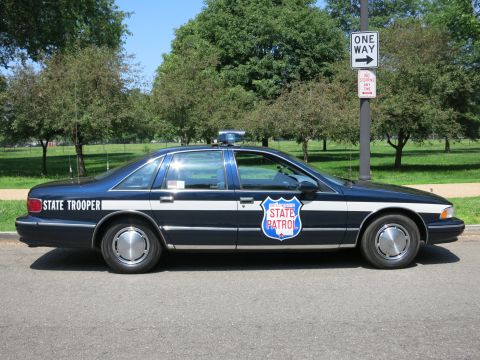 1991 Ford LTD Crown Victoria - dark blue and white State Trooper State Patrol sedan with a red shield decal parked near a One Way sign in a park.