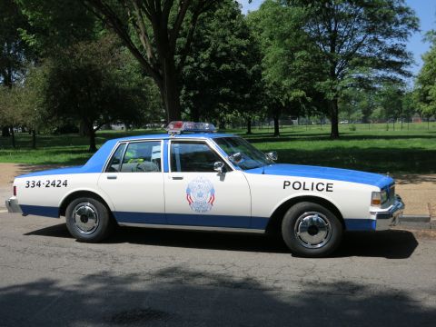 1991 Ford LTD Crown Victoria - blue and white police sedan marked Police and 334-2424 with a roof beacon parked in profile on a park road.