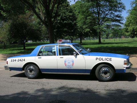 1991 Ford LTD Crown Victoria - Profile view of a blue and white police sedan lettered Police and 334-2424 with a circular emblem parked under trees.