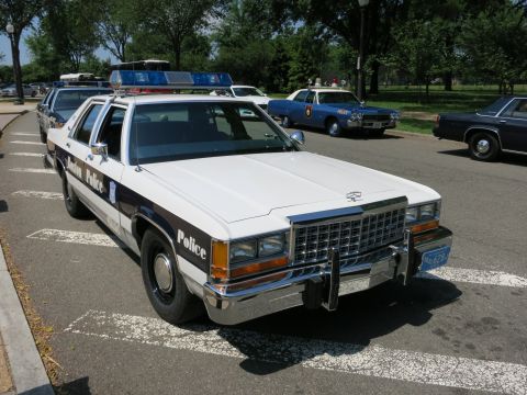1991 Ford LTD Crown Victoria - white and dark Ford police sedan lettered Police with a front push bar and roof lightbar parked among other cruisers.