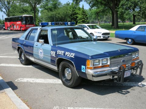 1991 Ford LTD Crown Victoria - two-tone blue Ford State Police sedan marked 90E with a push bar and circular seal parked among other patrol cars.