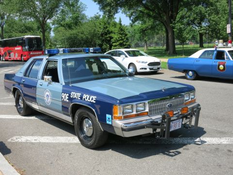 1991 Ford LTD Crown Victoria - Front three-quarter view of a two-tone blue State Police Ford sedan marked 90E with a push bar parked among cruisers.