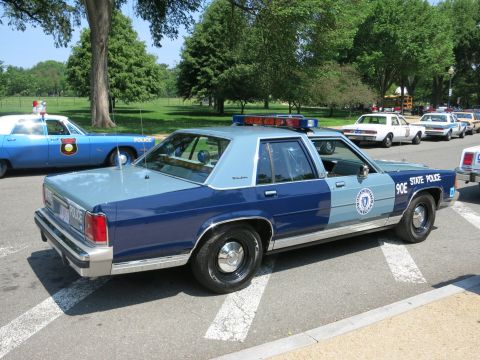 1991 Ford LTD Crown Victoria - Rear quarter view of a two-tone blue State Police Ford sedan marked 90E parked in a lot among other vintage cruisers.