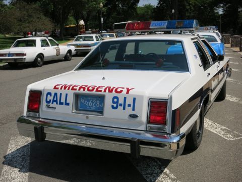 1991 Ford LTD Crown Victoria - Rear view of a white police sedan lettered Emergency Call 9-1-1 with a roof lightbar parked among vintage cruisers.
