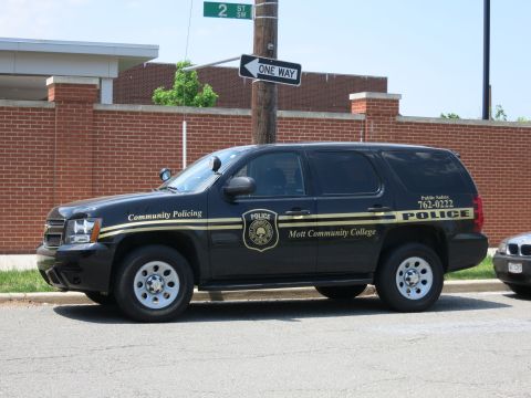 1991 Ford LTD Crown Victoria - black Chevrolet SUV marked Mott Community College Police with gold striping parked along a curb by a brick wall.
