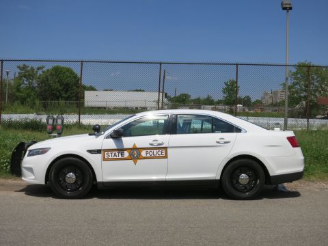 1991 Ford LTD Crown Victoria - Side profile of a white Ford State Police sedan with an orange door shield parked by a chain-link fence and field.