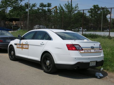 1991 Ford LTD Crown Victoria - Rear three-quarter view of a white Ford State Police sedan with orange door shield and plate 15-33 by a fence.