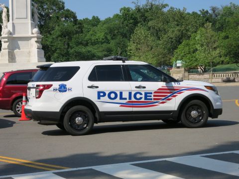 1991 Ford LTD Crown Victoria - white Ford Explorer police SUV marked POLICE with red-white-blue swoosh striping, parked near a statue and red minivan.